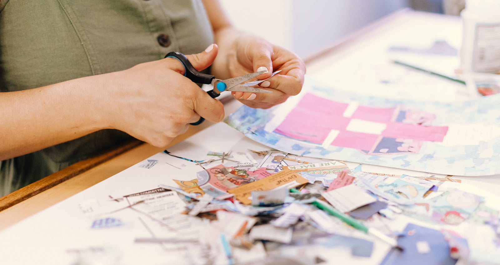 Bits of colorful newspaper are strewn around a work-in-progress collage on a clean white studio table. Catherine Elizabeth's hands hold a bit of newspaper in one hand and scissors in the other, diligently trimming pieces to add to the new collage.