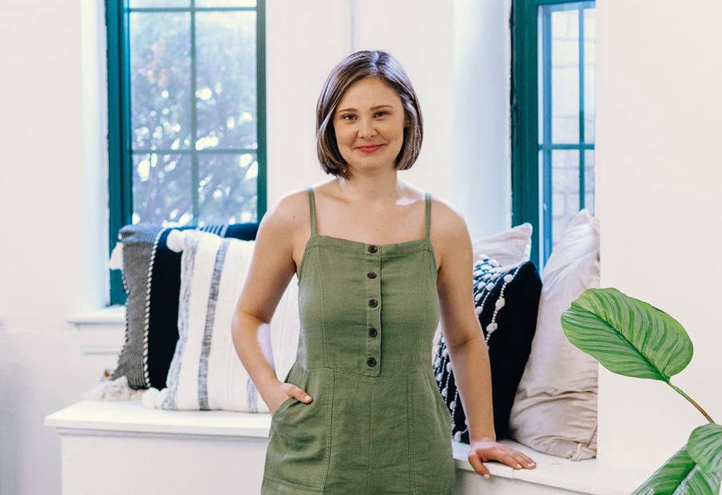 Catherine Elizabeth, a young white woman with short, neatly kept brown hair and a kind smile, stands near bright windows and soft black and white pillows in her studio space. She is wearing a summery green jumper with small buttons down the front.